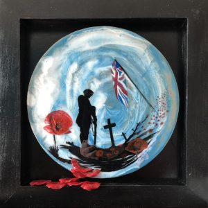 Black frame surrounding a circular painting with resin and acrylic depicting a British soldier bowing his head. Poppies and crosses dappled across the landscape with a flag waving in the wind. Artificial poppies across the front